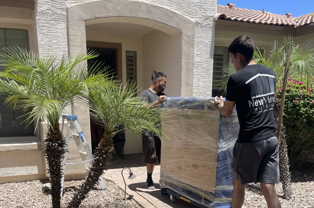 Two NewView movers wheel a large crated item past palm trees at a front door